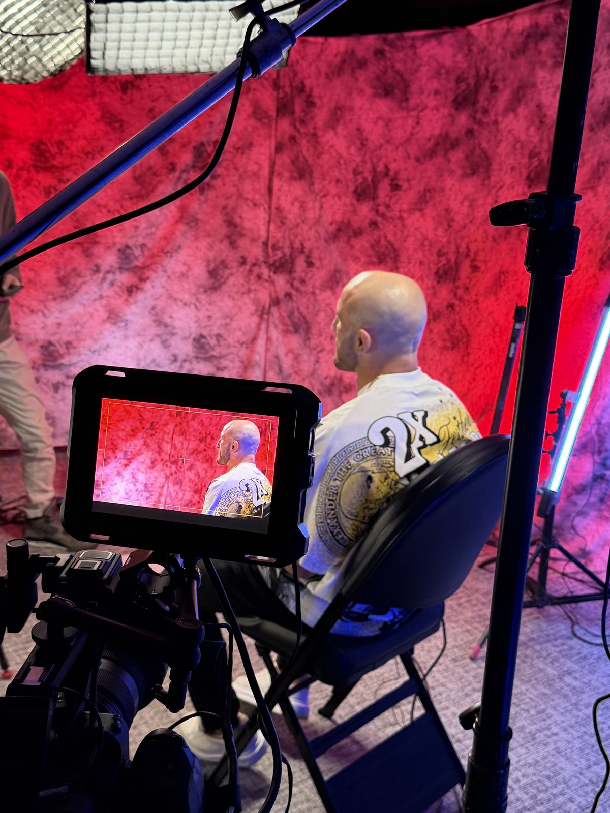 A man being filmed in front of a red backdrop, seen on a camera monitor