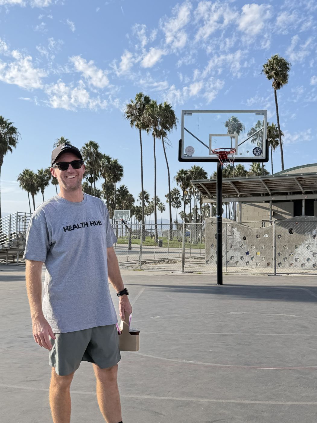 Mick Baines standing on the Venice Beach basketball courts with palm trees behind him, a drink in his hand