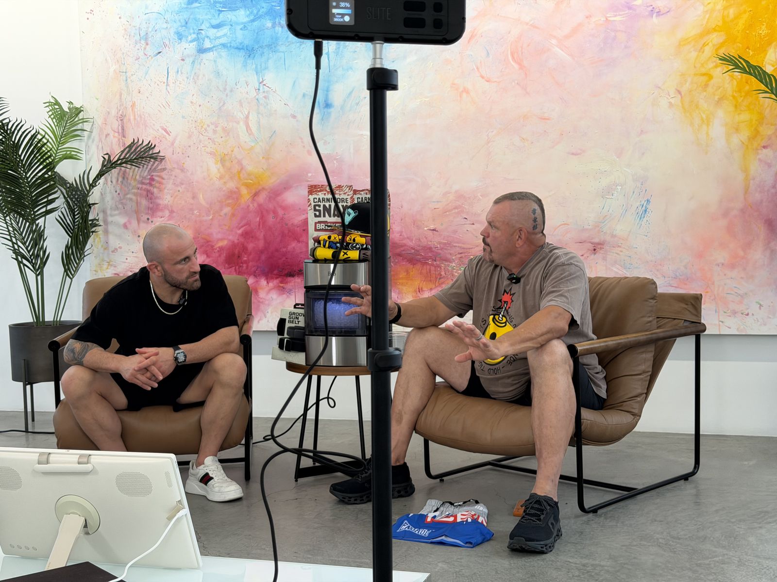 Alex Volkanovski and Chuck Liddell seated in leather chairs recording a podcast in front of a pink, yellow and blue painted wall