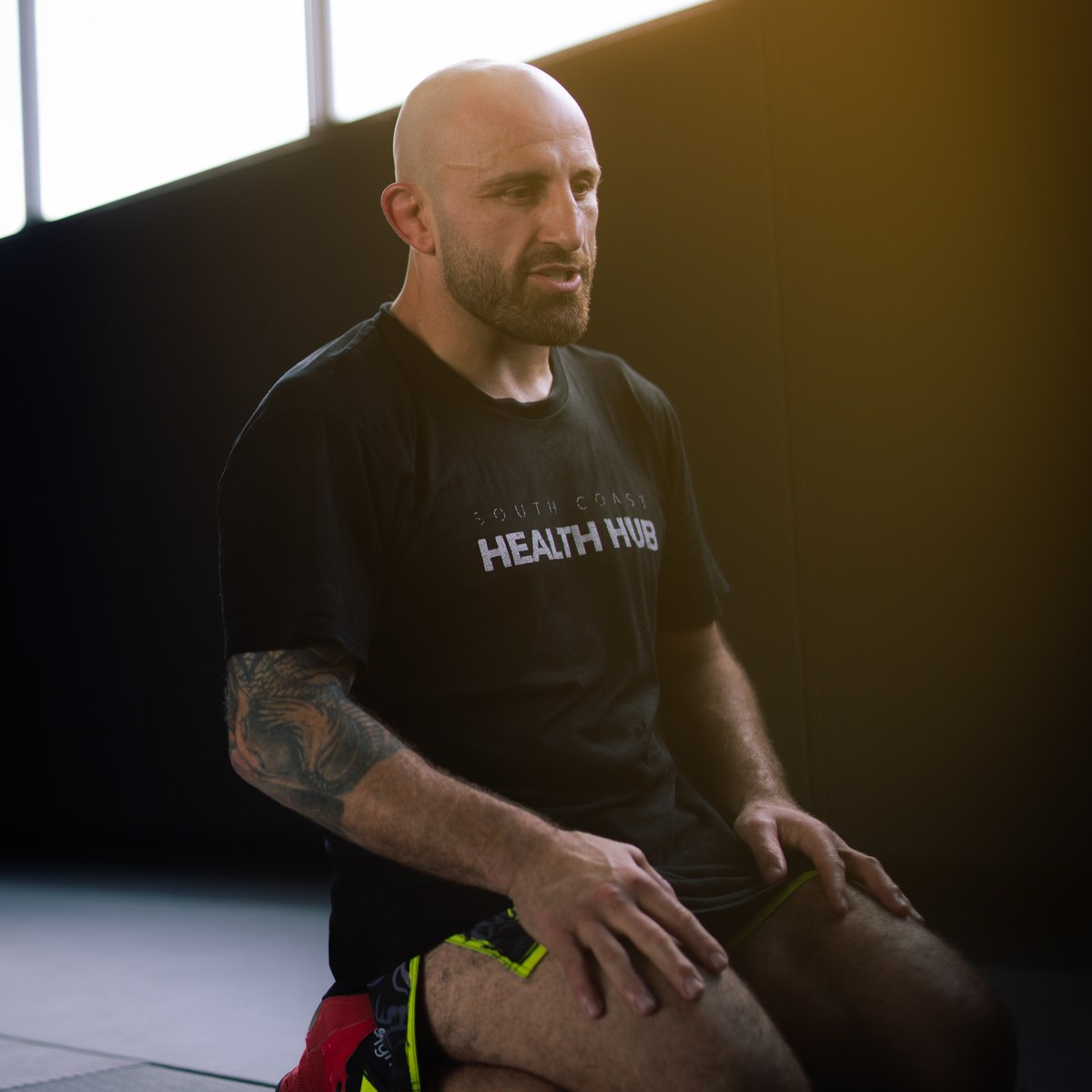 A man sitting on the mats stretching, in a South Coast Health Hub shirt