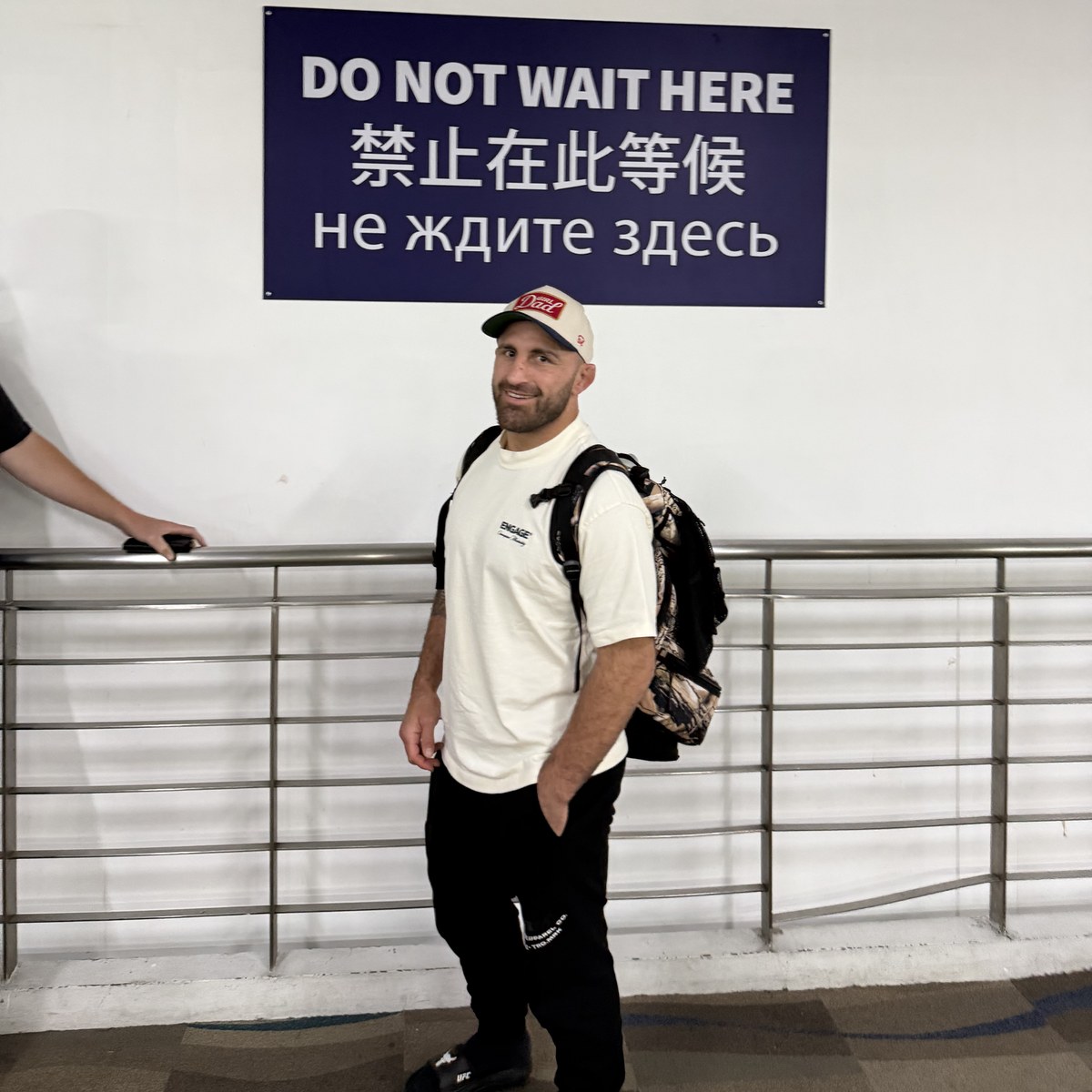 A man with a backpack standing under a trilingual sign reading Do Not Wait Here