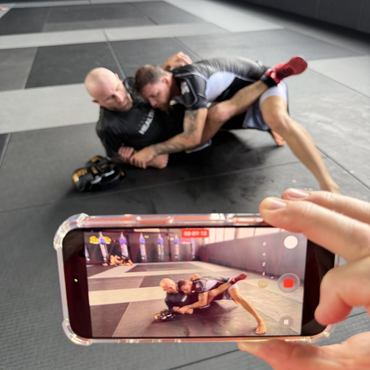 Two men grappling on the mats, filmed on a phone held up in the foreground